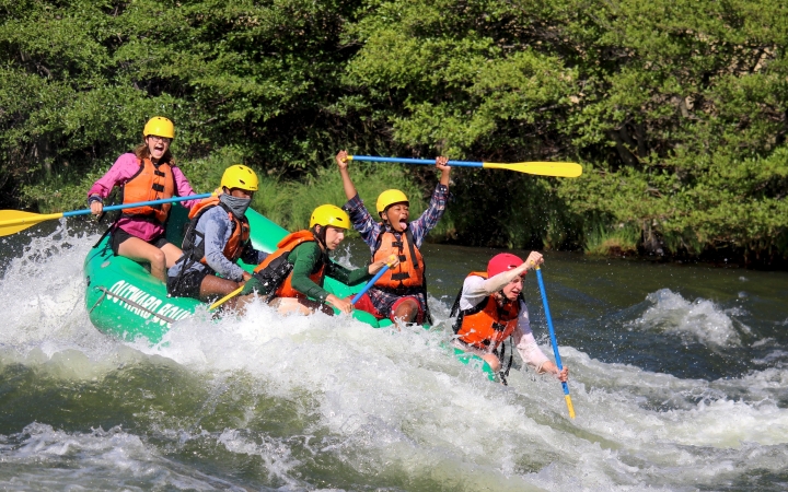 A group of students wearing safety gear paddle a raft through whitewater. One of them lifts their paddle into the air in apparent joy.